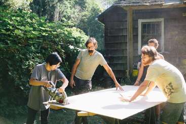 Four people are working together outdoors, cutting a large wooden board with a circular saw on saw horses, surrounded by greenery and an old wooden building.