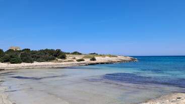 Uma cena serena à beira-mar mostra águas claras e rasas que se encontram com uma praia de areia. Dão-se a ver arbustos esparsos e um terreno rochoso, com um pequeno edifício de pedra ao longe.