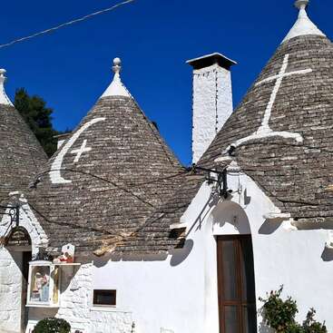 Duas casas tradicionais trulli, com telhados cônicos de pedra, exibem símbolos pintados de branco. O céu azul brilhante contrasta com as paredes brancas, criando um cenário arquitetônico pitoresco e único.
