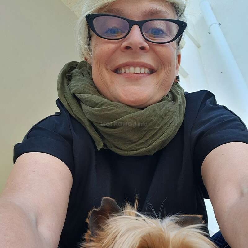 Uma mulher de óculos, com um lenço verde-oliva e uma blusa preta, sorri calorosamente para a câmera. A cabeça de um cachorrinho aparece na parte de baixo.