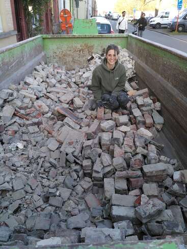 A woman sits in the back of a green dumpster filled with rubble, surrounded by a street scene with cars and people in the background.