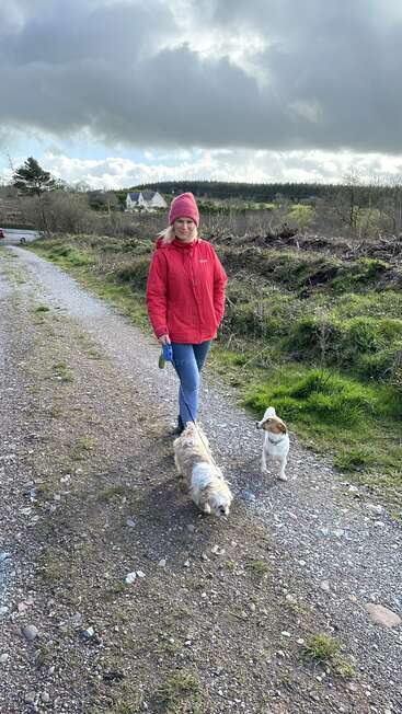 A woman walks two small dogs on a gravel path, wearing a red jacket and pink beanie, with houses and trees in the background under a cloudy sky.