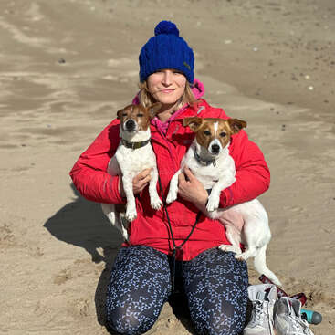 A woman sits on a sandy beach, cradling two small white dogs with brown ears and noses, wearing a blue beanie and red jacket.