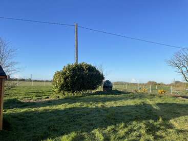 The image depicts a serene rural landscape featuring a lush green field, a large bush, a utility pole, and a small shed, set against a clear blue sky.