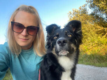 The image depicts a woman with blonde hair and sunglasses posing for a selfie with a black and white dog on a gravel path surrounded by trees and grass.