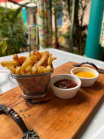 Auf einem Holztablett liegen knusprige Pommes frites in einem Metallkorb, begleitet von zwei Schalen mit Ketchup und Käsesoße, in einer charmanten Umgebung im Freien.