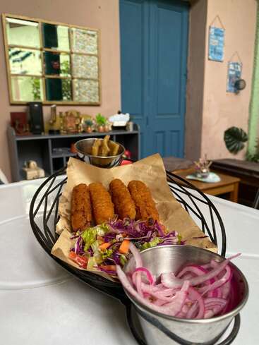 Ein Metallkorb enthält knusprig gebratene Spieße, frischen gemischten Salat und eingelegte rote Zwiebeln. Im Hintergrund ist ein gemütlicher Raum mit blauem Dekor und Wandkunst zu sehen.