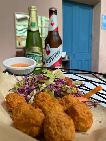 Eine Nahaufnahme von knusprig gebratenen Vorspeisen, frischem Salat, Dip-Sauce und zwei kalten Bieren (Suprema, Pilsener) auf einem Tisch in entspannter Umgebung.