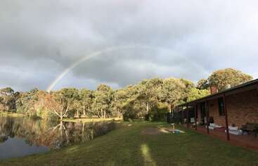 Um arco-íris se estende sobre uma paisagem serena com árvores, um lago que reflete a luz e uma casa de tijolos sob um céu nublado, criando uma cena rural tranquila e pitoresca.