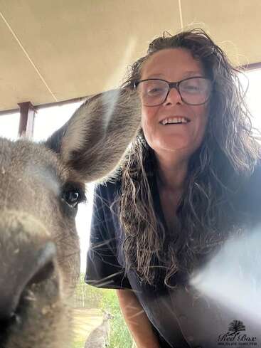 A smiling woman with long, wavy hair and glasses poses for a selfie with a curious kangaroo, while another kangaroo stands in the background outdoors.