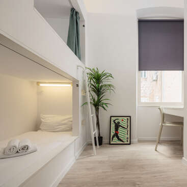 Une chambre moderne et minimaliste comprend des lits superposés, un bureau blanc, une plante verte, des œuvres d'art encadrées, un parquet clair et une fenêtre avec un store roulant foncé.