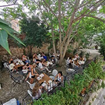 L'image représente une salle à manger extérieure sereine, où de nombreuses personnes sont assises à des tables sous un arbre majestueux, entouré d'une végétation luxuriante et d'un mur de pierre.