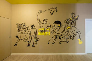 Une fresque murale représente des dessins stylisés au trait noir représentant des personnes et un oiseau, rehaussés de touches de jaune vif, vaquant à leurs occupations quotidiennes sur un mur brun clair.