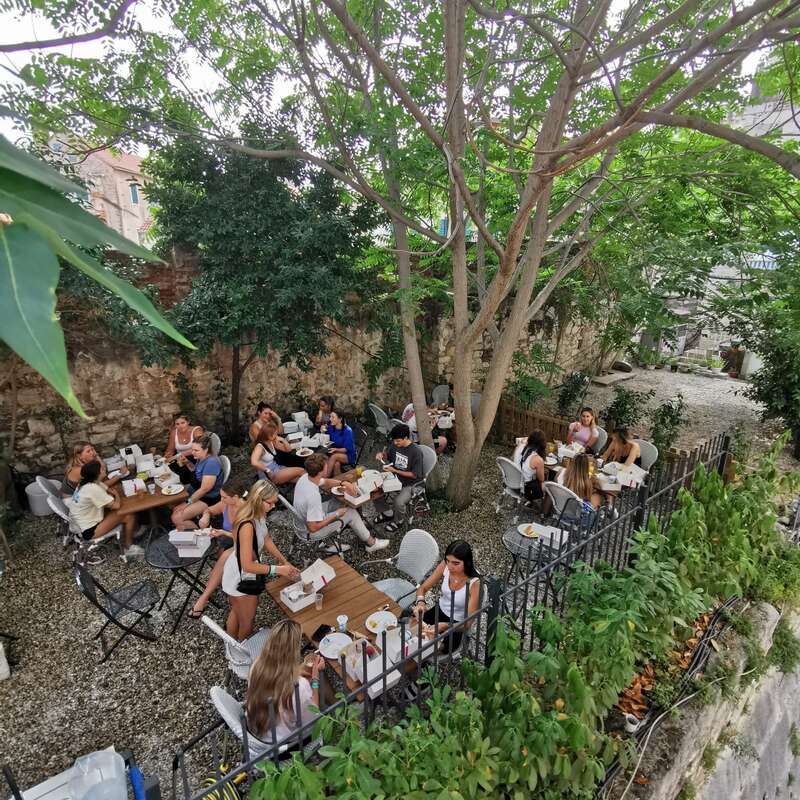 A imagem mostra uma área de jantar serena ao ar livre, onde várias pessoas estão sentadas em mesas embaixo de uma árvore majestosa, cercadas por uma vegetação exuberante e um muro de pedra.
