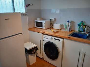 Essa pequena cozinha contém uma geladeira, máquina de lavar, micro-ondas, torradeira, material de limpeza, uma pia azul, uma lixeira e bancadas de madeira sob uma janela.