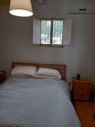 Um quarto aconchegante com cama de madeira, roupa de cama listrada, dois travesseiros, pequena mesa de cabeceira de madeira, abajur, janela com grades, ar-condicionado e paredes brancas; decoração simples.