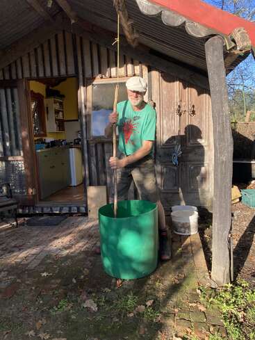 A man wearing a green t-shirt and cap stirs a large green container outdoors, beside a rustic wooden cabin with an open door and a bucket nearby.