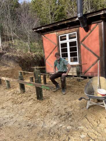 A man in muddy clothes sits on wooden beams in front of a small, rustic house. A wheelbarrow and construction materials are scattered around the area.