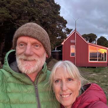 A smiling older couple in warm jackets stands outside a red cabin at sunset, surrounded by trees, enjoying a peaceful, nature-filled outdoor setting together.
