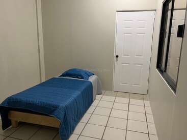 Esta imagen muestra un dormitorio sencillo y pequeño con paredes blancas, suelo de baldosas, una cama individual con ropa de cama azul, una puerta blanca y una ventana.