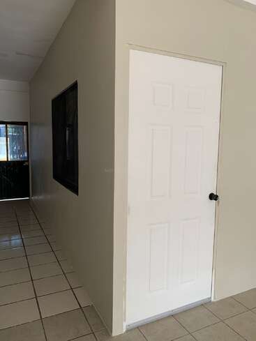 En un pasillo embaldosado hay una pequeña habitación de paredes beige, con una puerta blanca con un pomo negro y una gran ventana en un lado.