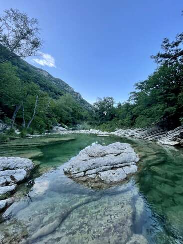 Un río tranquilo de aguas cristalinas y color esmeralda discurre entre lechos rocosos y frondosos árboles, con las montañas como telón de fondo bajo un cielo azul brillante.