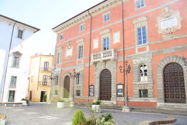 Una plaza europea bañada por el sol, con edificios históricos de fachadas coloridas, puertas con arcos, ventanas altas, mampostería decorativa, farolas, bancos y macetas con plantas en primer plano.