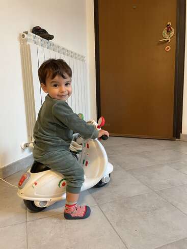 Un niño pequeño, sonriente, está sentado en un pequeño patinete de juguete blanco dentro de casa. Lleva un chándal verde y calcetines de colores. Hay un zapato apoyado sobre el radiador.