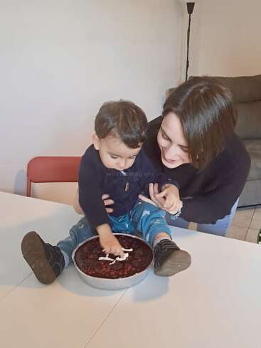 Un niño pequeño está sentado en una mesa con la mano metida en un pastel, mientras una mujer le sonríe y le sujeta con delicadeza. Ambos parecen felices, disfrutando juntos de un momento divertido.