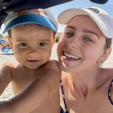 Una mujer sonriente y un niño pequeño se hacen un selfi en primer plano en una playa soleada de guijarros. Ambos llevan gorras y parecen felices, disfrutando del tiempo que pasan juntos.