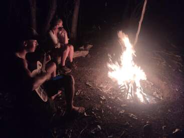 Deux personnes sont assises près d'un feu de camp, la nuit, dans les bois. L'un gratte une guitare tandis que l'autre surveille les flammes, créant ainsi une atmosphère confortable et paisible.