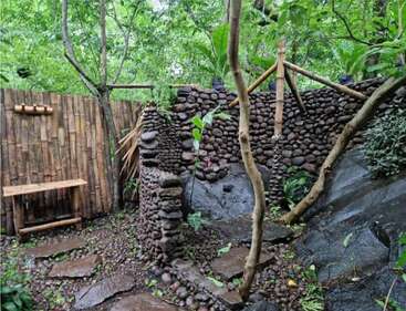Cette image représente une douche extérieure faite de pierres naturelles et de bambou, entourée d'une forêt verdoyante, se fondant harmonieusement dans l'environnement et évoquant une simplicité paisible.