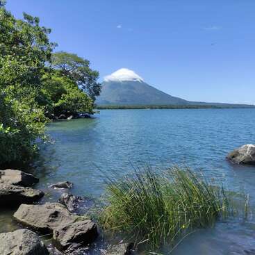 A tranquil lake with clear blue water, surrounded by lush greenery, rocks, and distant volcano. The volcano’s peak is partially covered by a fluffy white cloud.