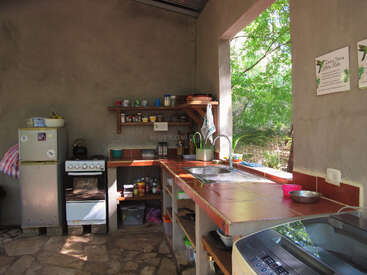 This image shows a rustic kitchen with stone floors, open shelves, a small fridge, stove, sink, and bright sunlight streaming through a window, highlighting outdoor greenery.