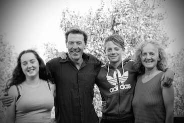 A black and white photo of four people standing close together, smiling. They appear to be a happy family, outdoors with trees in the background.