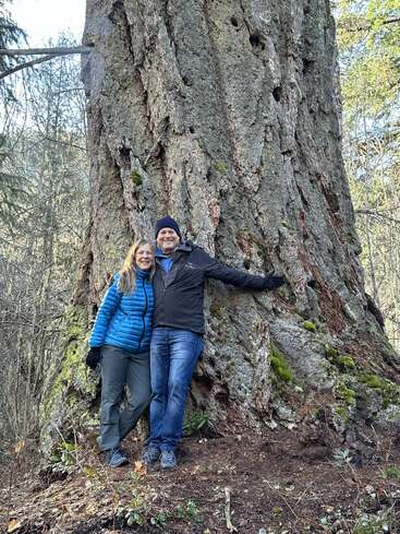Un couple souriant, vêtu de vêtements de plein air, se tient ensemble, adossé au tronc massif d'un arbre ancien dans une zone forestière, profitant de la nature.