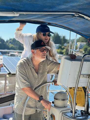 Un homme et une femme sont sur un bateau et naviguent. L'homme tient la barre tandis que la femme regarde devant elle. Tous deux portent un chapeau et des lunettes de soleil.