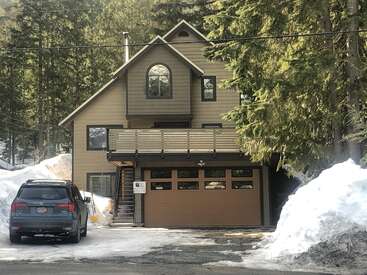Une maison moderne à deux étages avec un garage brun, partiellement entourée de neige et de grands pins. Un 4x4 noir est garé dans l'allée.
