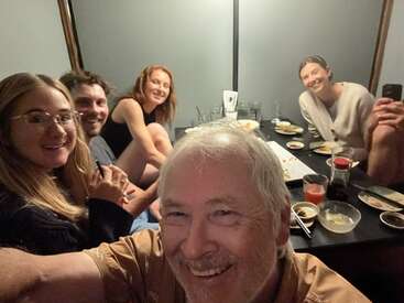 Un groupe de cinq personnes est assis autour d'une table, souriant et savourant un repas ensemble. L'atmosphère est chaleureuse, amicale et détendue.