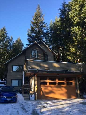A modern two-story house with a wooden garage, surrounded by tall evergreen trees and snow. A blue car is parked outside, under a clear sky.