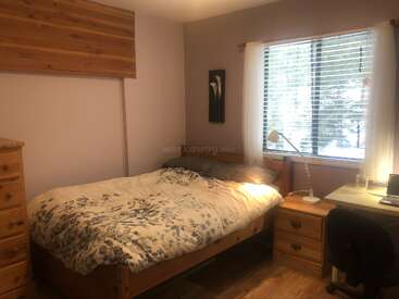 This cozy bedroom features a wooden bed, floral bedding, desk with lamp, dresser, and a large window with blinds, letting in plenty of natural light.