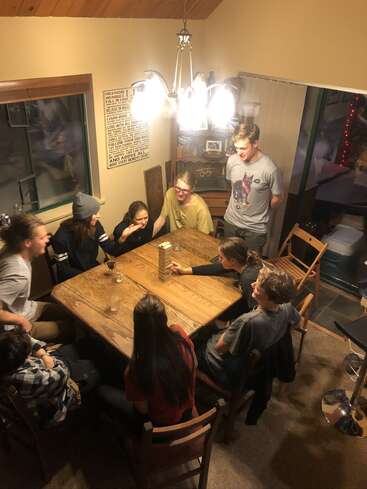 A group of friends gathers around a wooden table, focused on playing Jenga. Laughter, concentration, and cozy lighting create a warm, lively atmosphere in the room.
