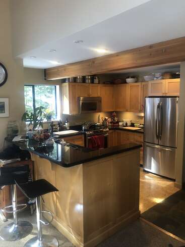 This kitchen features light wood cabinets, black countertops, stainless steel appliances, bar stools, and ample natural light from a large window; it's cozy, modern, and inviting.