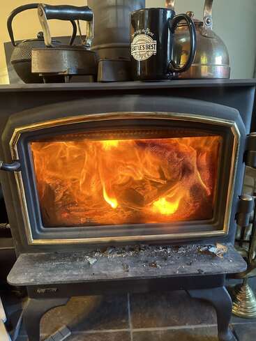 A cozy wood stove with bright flames burning inside. On top are a kettle, an iron, a teapot, and a Seattle’s Best Coffee mug. Warm ambiance.