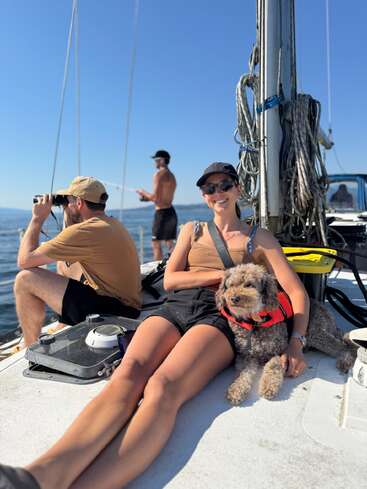 Three people and a dog relax on a sunny sailboat. One man uses binoculars, another stands shirtless, while a woman smiles beside her dog in a lifejacket.