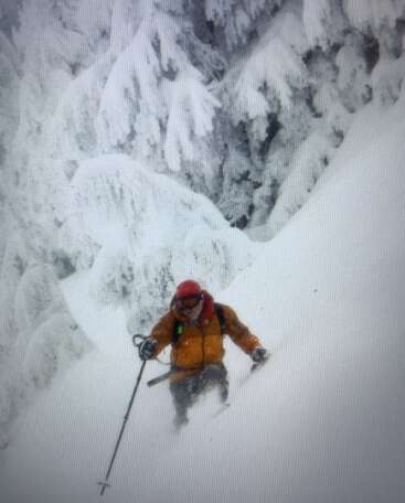 A skier in an orange jacket and red helmet navigates deep powder snow down a mountain, surrounded by heavily snow-laden evergreen trees in a winter wonderland.