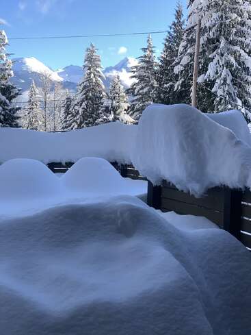 A thick blanket of snow covers a balcony, railing, and outdoor furniture. Snow-capped pine trees and distant mountains complete this beautiful, serene winter landscape.
