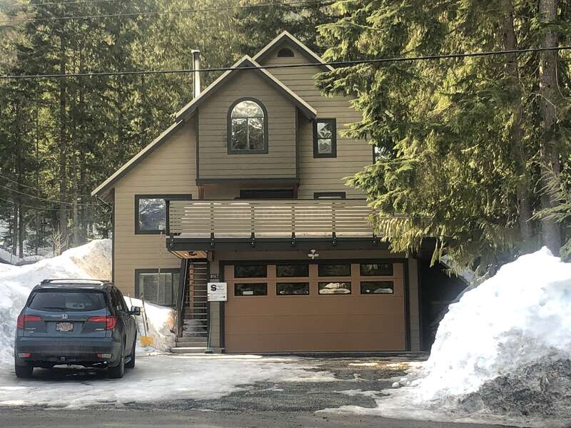 Une maison moderne à deux étages avec un garage brun, partiellement entourée de neige et de grands pins. Un 4x4 noir est garé dans l'allée.