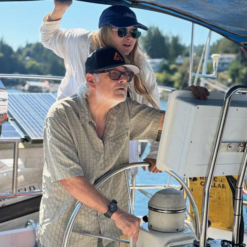 A man and woman are on a boat, navigating. The man is steering the wheel while the woman looks ahead. Both wear hats and sunglasses.