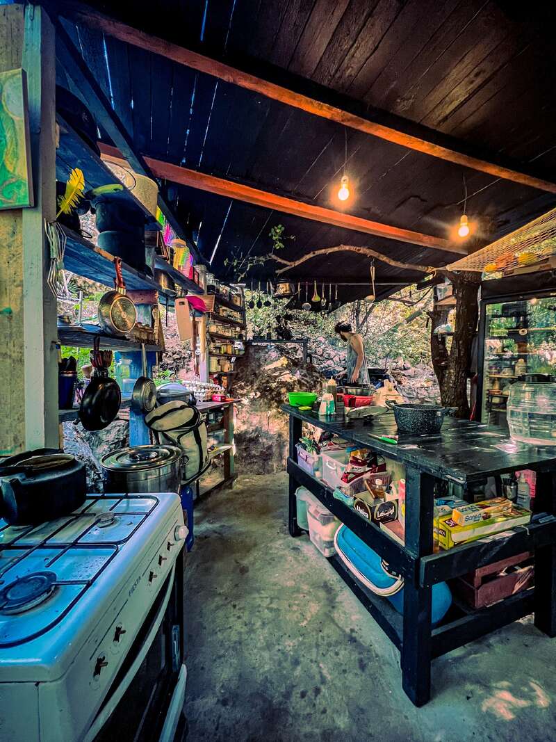 A rustic, open-air kitchen with shelves full of utensils and supplies, a gas stove, wooden beams, warm lighting, and natural surroundings, creating a cozy, creative atmosphere.